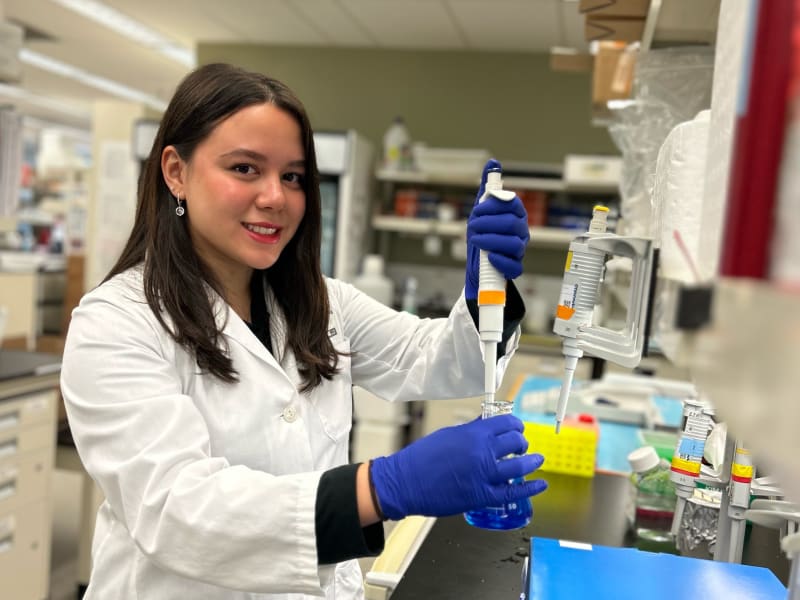 A women with brown hair wearing a white lab coat smiles for the camera while mixing research samples in a beaker. 